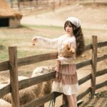A young woman in vintage attire feeds sheep on a sunny farm, offering a rustic countryside charm.